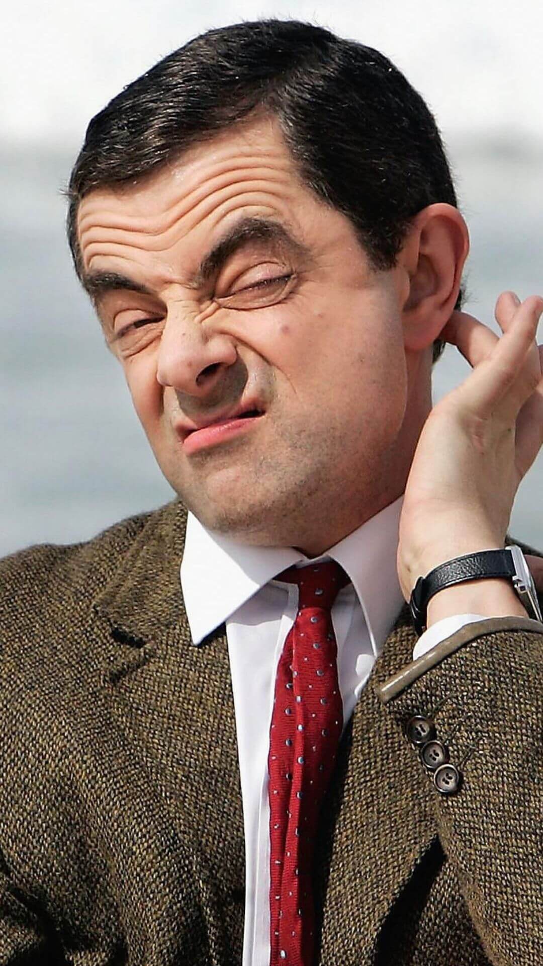 mr bean in a brown coat and red tie making a funny face