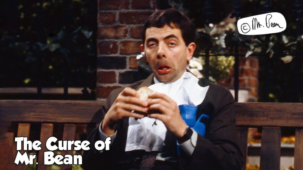 mr bean sitting on a bench in a park about to sneeze